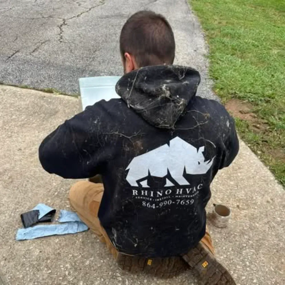 A Rhino HVAC technician kneeling in a Simpsonville driveway to repair an equipment panel