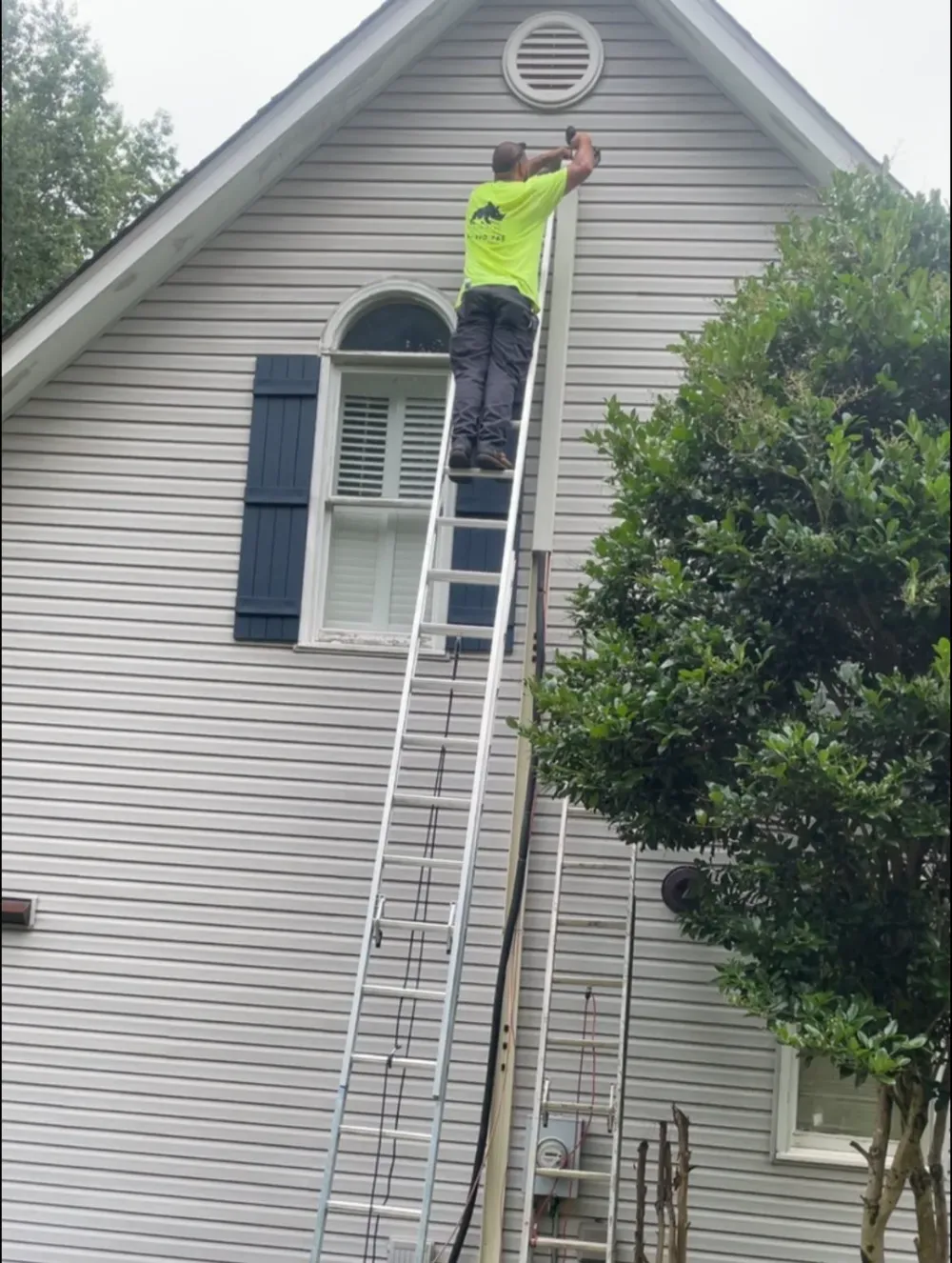 A Rhino HVAC technician on a ladder at the roofline of a Simpsonville home, working on a vent